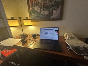 My workspace at the La Quinta Inn. My laptop is on the provided desk, a few pages of printed manuscript are to the left of the laptop along with a book, a morass of charging cables and a HomePod mini. To the right of the laptop is my iPad and above the laptop is a generic corporate art painting on the wall.